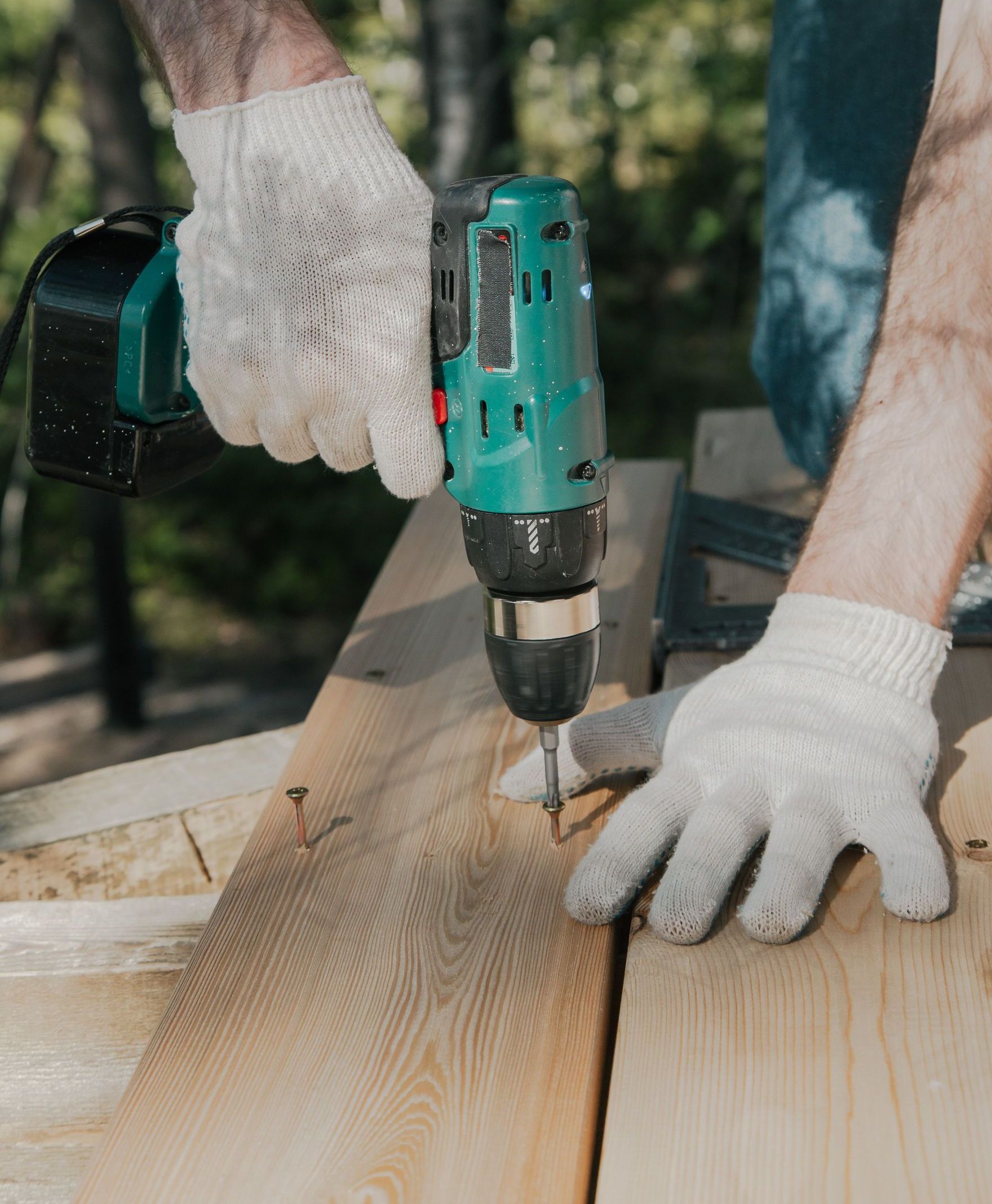 Adult using a power drill on a wooden surface, wearing white gloves, outdoors in daylight.